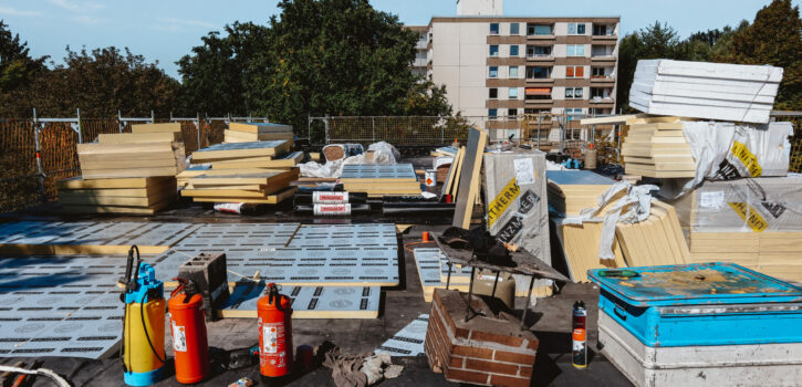 Das Bild zeigt den Einbau von Gefälledämmeinlagen während einer Flachdachsanierung in der Nähe von Düsseldorf. Die Fotoaufnahme zeigt zwei Dachdecker, welche die Bitumenabdichtungslagen an der Attika sowie an einem Technikhäuschen anarbeiten. Die KKOe Bausachverständige und Ingenieure war mit der Bauleitung beuauftragt.