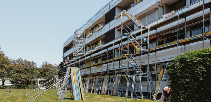 Das Bild zeigt ein stellenweise fertig aufgebautes Gerüst an einer Fassade eines Mehrfamilienhauses in der Nähe von Düsseldorf, dessen Balkonbrüstungen saniert werden sollen. Die Fotoaufnahme zeigt zwei Dachdecker, welche die Bitumenabdichtungslagen an der Attika sowie an einem Technikhäuschen anarbeiten. Die KKOe Bausachverständige und Ingenieure war mit der Bauleitung beuauftragt.