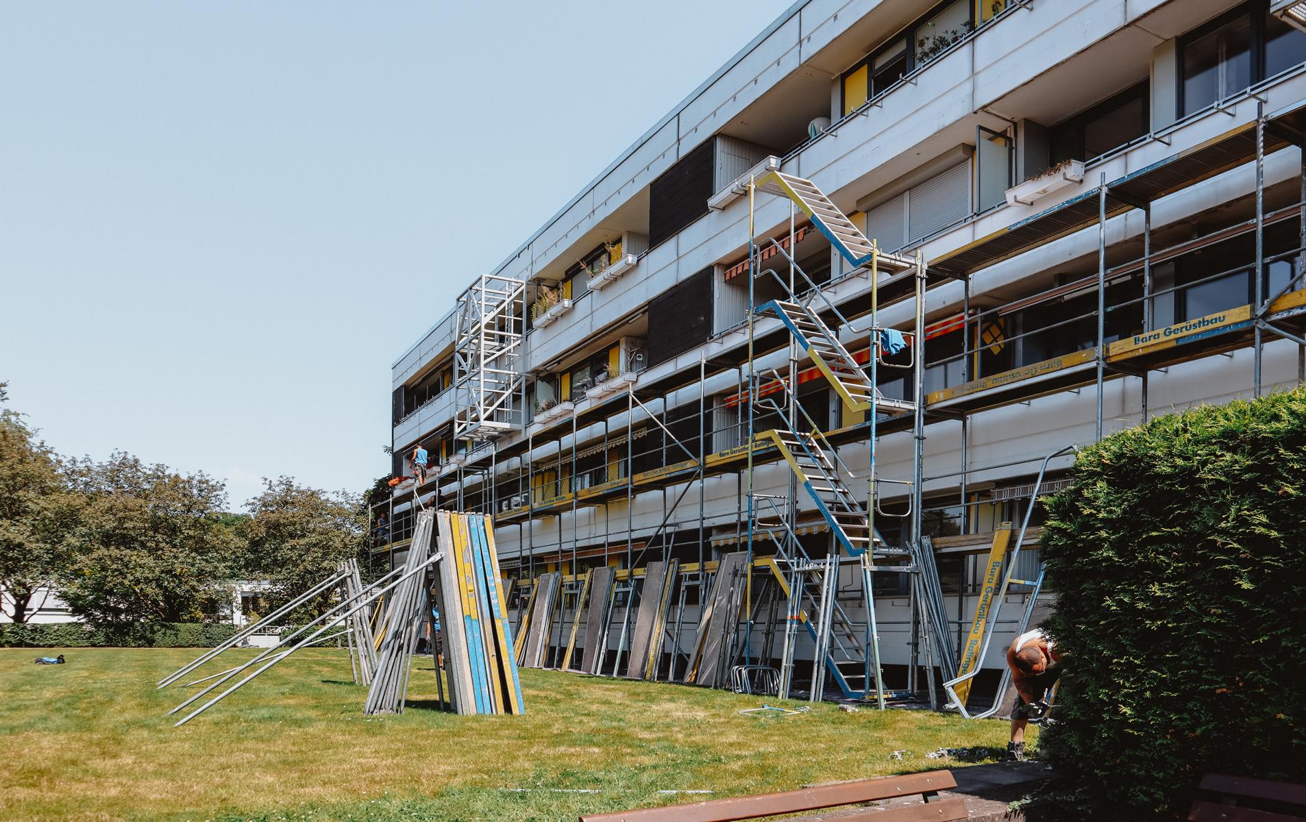 Das Bild zeigt ein stellenweise fertig aufgebautes Gerüst an einer Fassade eines Mehrfamilienhauses in der Nähe von Düsseldorf, dessen Balkonbrüstungen saniert werden sollen. Die Fotoaufnahme zeigt zwei Dachdecker, welche die Bitumenabdichtungslagen an der Attika sowie an einem Technikhäuschen anarbeiten. Die KKOe Bausachverständige und Ingenieure war mit der Bauleitung beuauftragt.