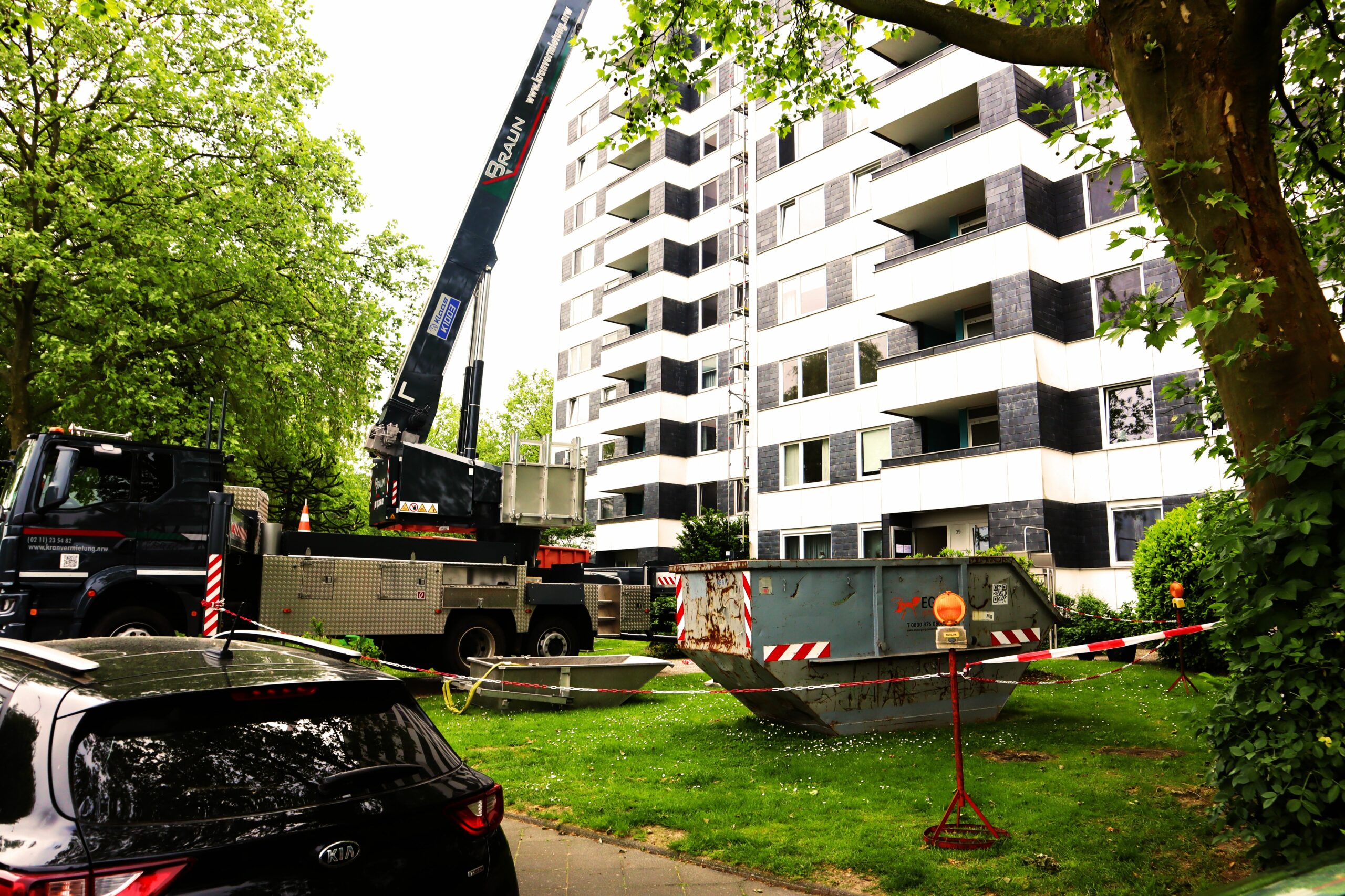 Das Bild zeigt einen Kraneinsatz im Zuge von Abbrucharbeiten an einem Flachdach in der Nähe von Düsseldorf. Die Fotoaufnahme zeigt zwei Dachdecker, welche die Bitumenabdichtungslagen an der Attika sowie an einem Technikhäuschen anarbeiten. Die KKOe Bausachverständige und Ingenieure war mit der Bauleitung beuauftragt.