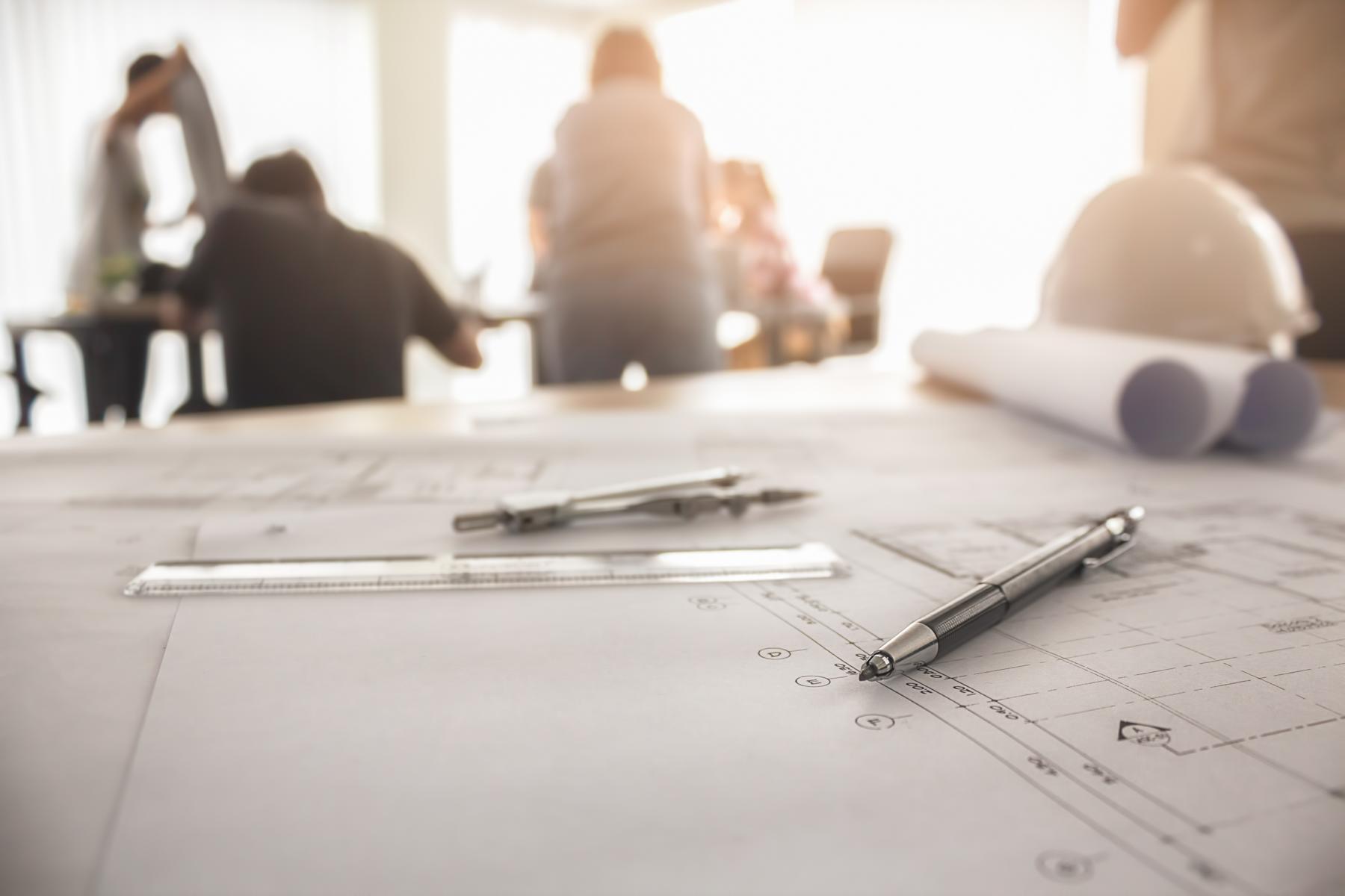 Das Bild zeigt im Hintergrund eine Gruppe von Ingenieuren, die an einem Meeting im Rahmen einer Bauplanung teilnehmen. Im Vordergrund des Bilds ist eine Grundriss auf einer Planzeichnung zu erkennen.