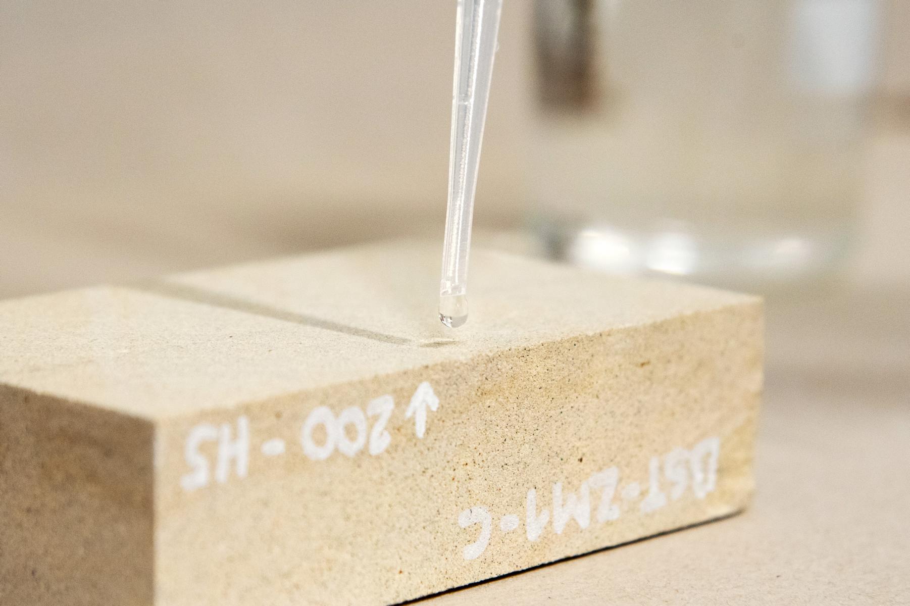 Prüfung der Wirksamkeit einer Hydrophobierung auf einem Sandstein Das Bild zeigt eine mit destilliertem Wasser gefüllte Pipette, an deren Spitze sich eine Wassertropfen gebildet hat, der zeitnah auf einen darunter gelegenen (formatierten) Sandstein tropfen wird. Wenn der Tropfen von dem Sandstein aufgesogen wird, ist die Hydrophobierung wirkungslos; sollte der Tropfen hingegen auf der Sandsteinoberfläche "stehen" bleiben, beweist das die Wirksamkeit der Hydrophobierung.