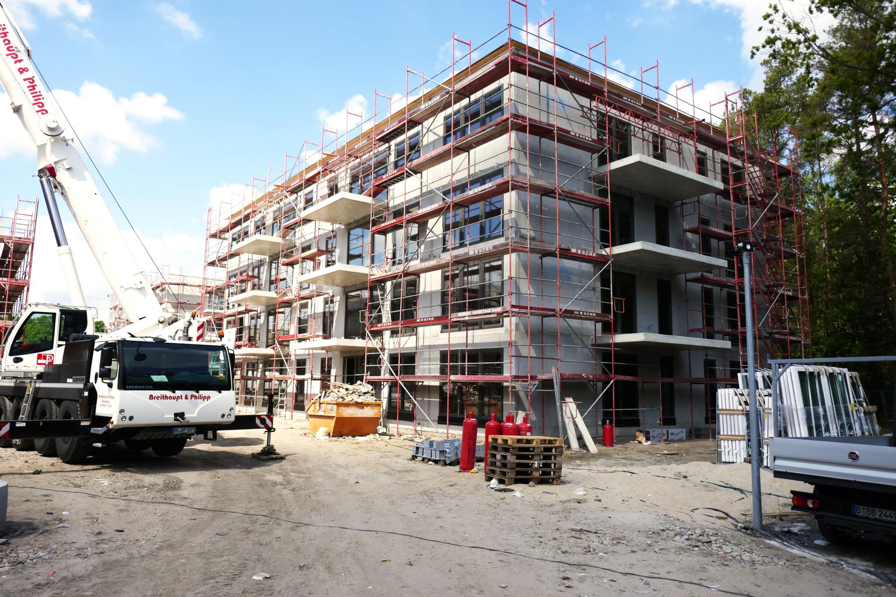 Das Bild zeigt einen Rohbau, um den ein Gerüst (zwecks Arbeiten an der Fassade) gestellt wurde. Die Fensterelemente sind bereits eingebaut worden. Des Weiteren steht neben dem Gebäude ein mobiler Kran, mit dessen Hilfe Material auf das Flachdach des Rohbaus transportiert wird.