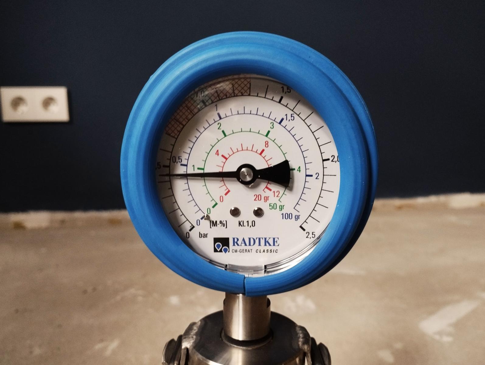 Das Bild zeigt das Manometer der Prüfflasche, in der sich vier Stahlkugeln, eine Calciumcarbid-Ampulle sowie Probenmaterial befindet. Durch das Schütteln der Prüfflasche, zerbricht die Calciumcarbid-Ampulle aufgrund der herumwirbelnden Kugeln. Das Calciumcarbid reagiert dann mit dem feuchten Probenmaterial. Es entsteht Acetylen (ein Gas), was dazu führt, dass der Druck in der Prüfflasche steigt. Dieser Druck wird kann am Manometer abgelesen werden und ist ein Maß für den Wassergehalt der Probe.