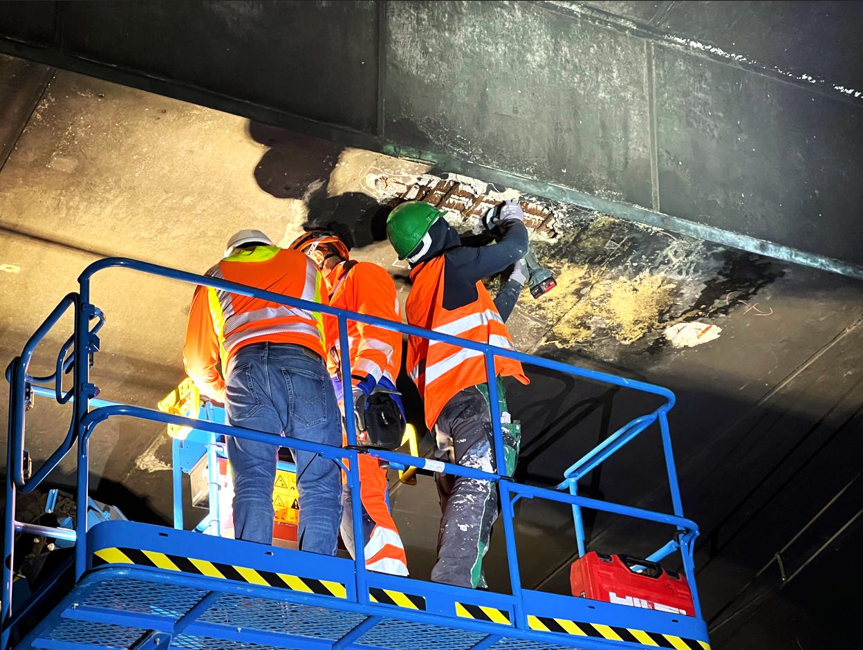 Das Bild zeigt die nicht zerstörungsfreie Bauwerksprüfung im Rahmen einer Bauwerksinspektion an einem Brückenbauwerk, welches mit Spannbetonbauteilen konstruiert wurde. Im Rahmen der Untersuchung wurde der Beton im Bereich der Hüllrohre freigelegt; anschließend wurden diese geöffnet, um den Einpressmörtels der Stahllitzen zu beproben. Der entnommene Einpressmörtels wurde seinen Chlorid- und Nitrat sowie Sulfatgehalt geprüft, um die Standfestigkeit des Brückenbauwerks zu beurteilen.