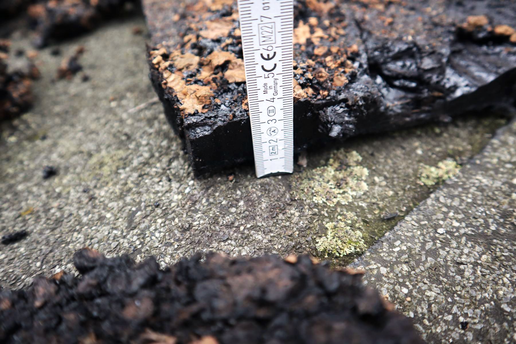 Das Bild zeigt ein Stück einer Bitumenbahn, welche im Zuge einer Bauteilöffnung aus einem Flachdachaufbau entfernt wurde. Die Dicke der Bitumenbahn wurde mithilfe eines "Zollstocks" gemessen. Die Information ist für die Planung einer fachgerechten Sanierung enorm wichtig.